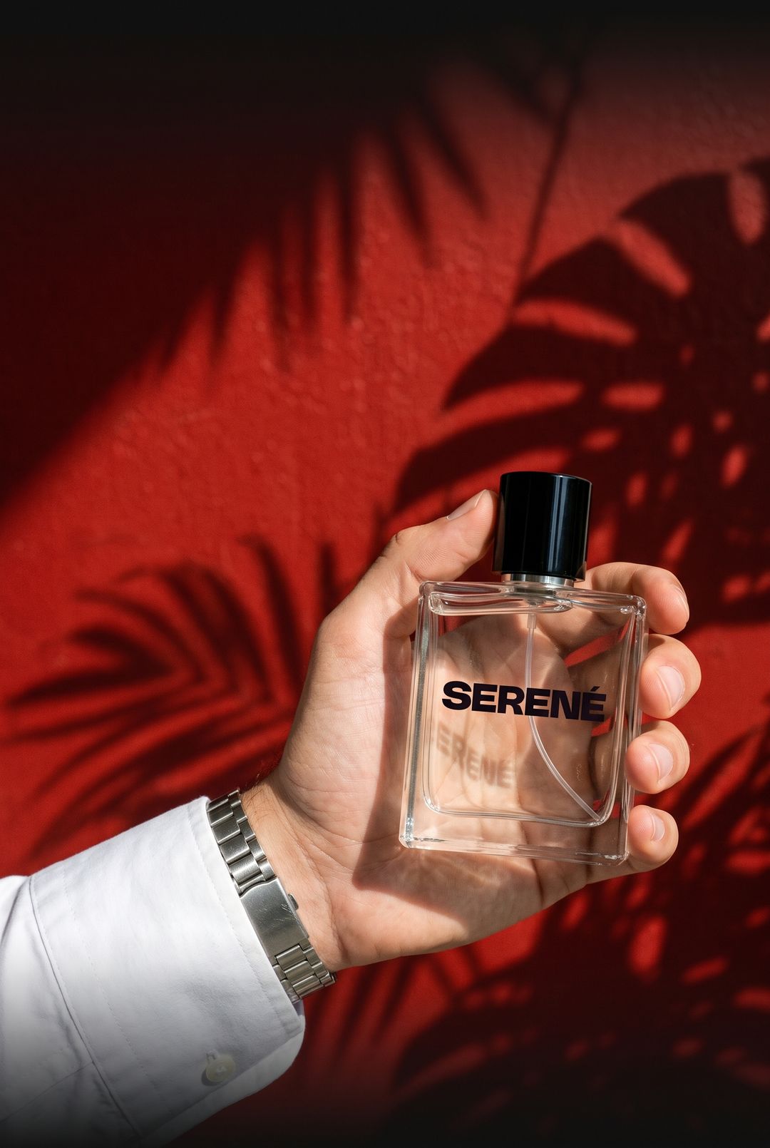 Mano con camisa blanca y reloj sosteniendo un perfume Serené frente a una pared roja al sol